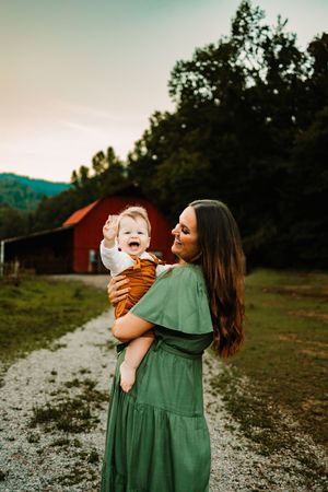 A smiling mother in a green dress holds her laughing baby near a red barn at sunset.