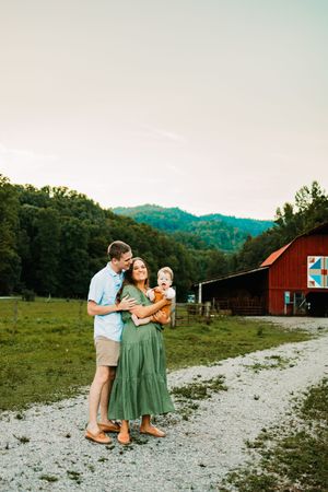 A happy family poses on a gravel path by a red barn, mother in green dress holding baby.