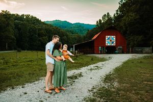 Young family with toddler posing near a red barn on a scenic farm at sunset.