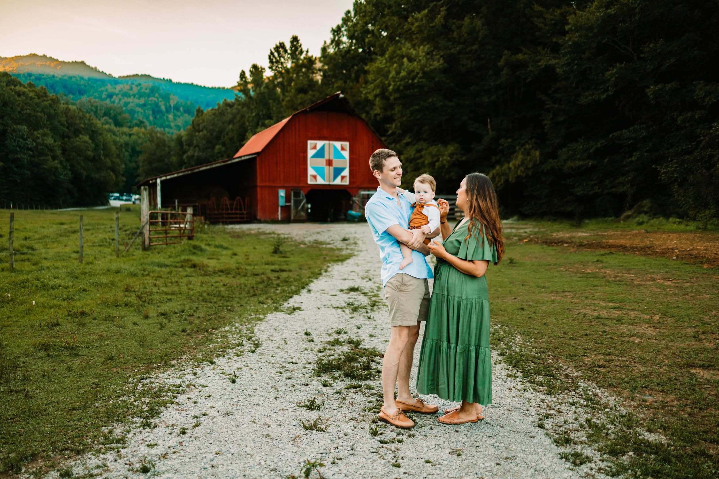 Family of three poses on gravel path before red barn with quilt square, green mountains backdrop at golden hour.