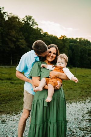 Happy couple with baby at sunset, man kissing woman in green dress holding child in orange outfit.