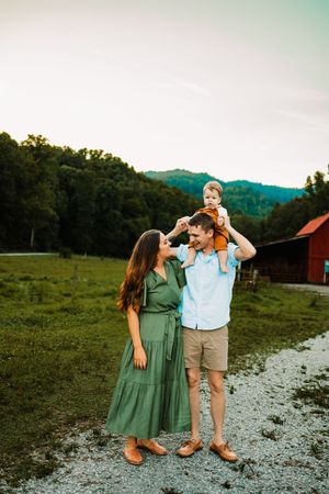 Family of three outdoors, baby sitting on fathers shoulders while mother looks up smiling near red barn.