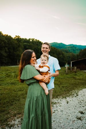 A young family poses outdoors at golden hour, mother in green dress holding baby in rust outfit with smiling father.