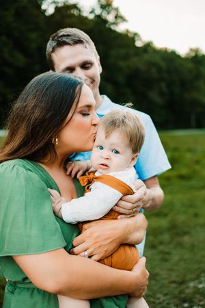 Mother kisses blue-eyed baby held by father in green field at sunset.