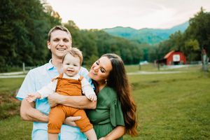 Happy family with baby in orange outfit posing outdoors with mountain and red barn backdrop.