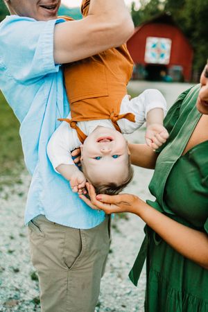 Father playfully holds baby upside down while mother laughs on rural farm.