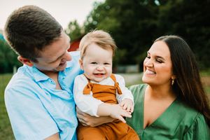 Happy young family with smiling baby in orange overalls outdoors, parents laughing together in a warm family portrait.