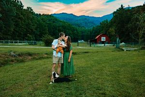 Couple kisses while holding baby with mountain and red barn backdrop at dusk.