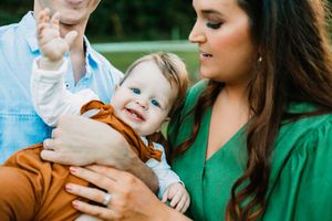 Close-up of happy blue-eyed baby in rust romper smiling while being held by parents in green and blue outfits.