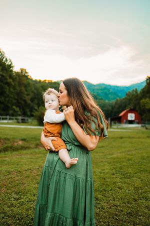 Mother in green dress kissing her baby while standing in a scenic green field near mountains.