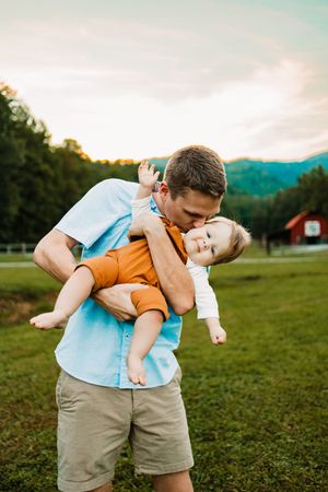 Father kissing baby outdoors in green field with mountains and red barn in background at sunset.