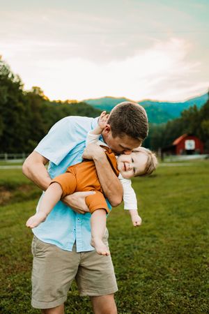 Father in light blue shirt playfully holds laughing baby in rust outfit against scenic mountain backdrop.