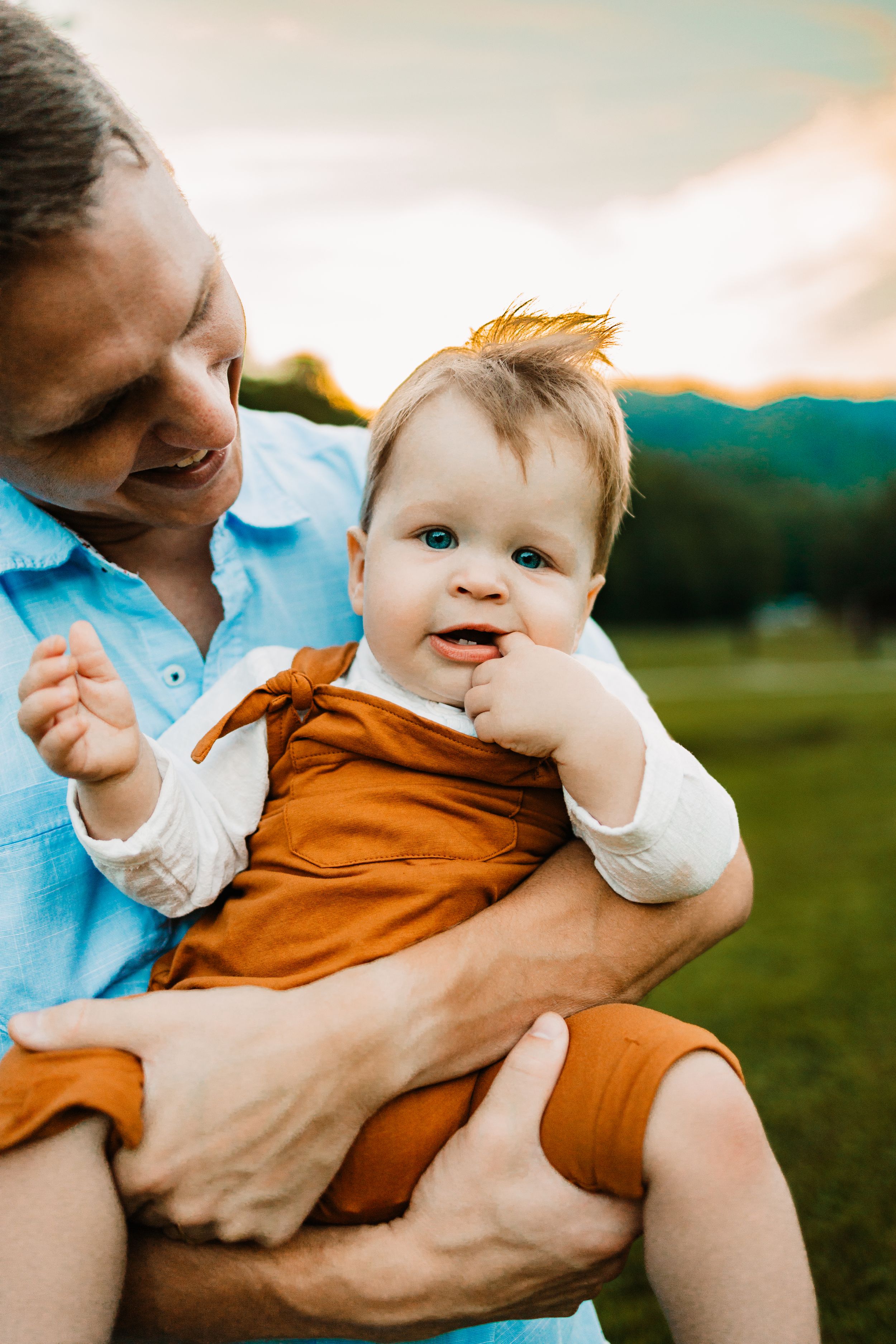A smiling man holds a happy blue-eyed baby in an orange outfit outdoors with green hills at sunset.