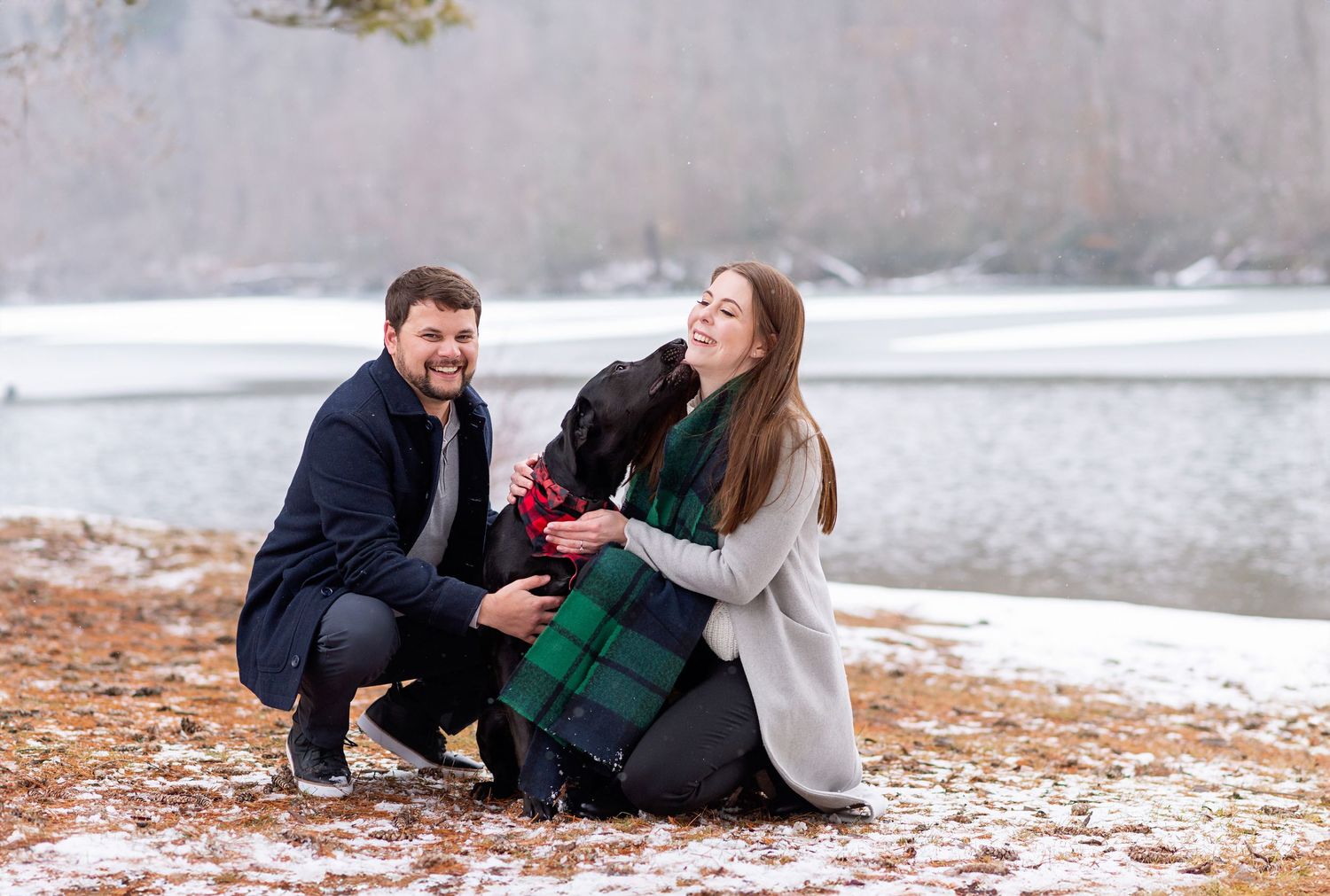 Couple crouching outdoors in snow with black Labrador dog wearing plaid scarf, smiling by a winter lake.