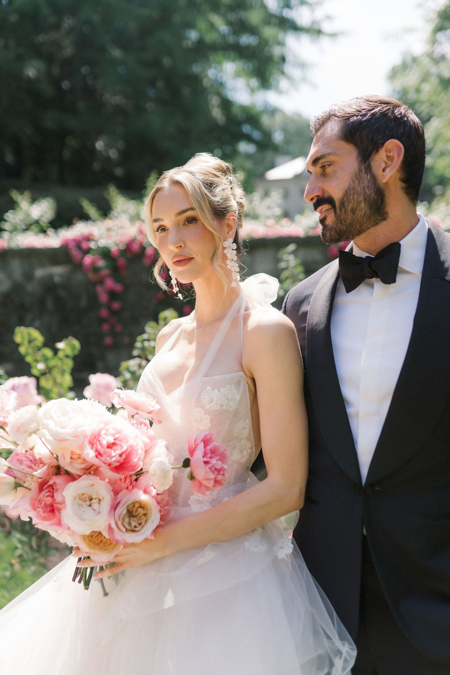 Bride holding pink peony bouquet in white gown beside groom in black tuxedo outdoors in garden setting.