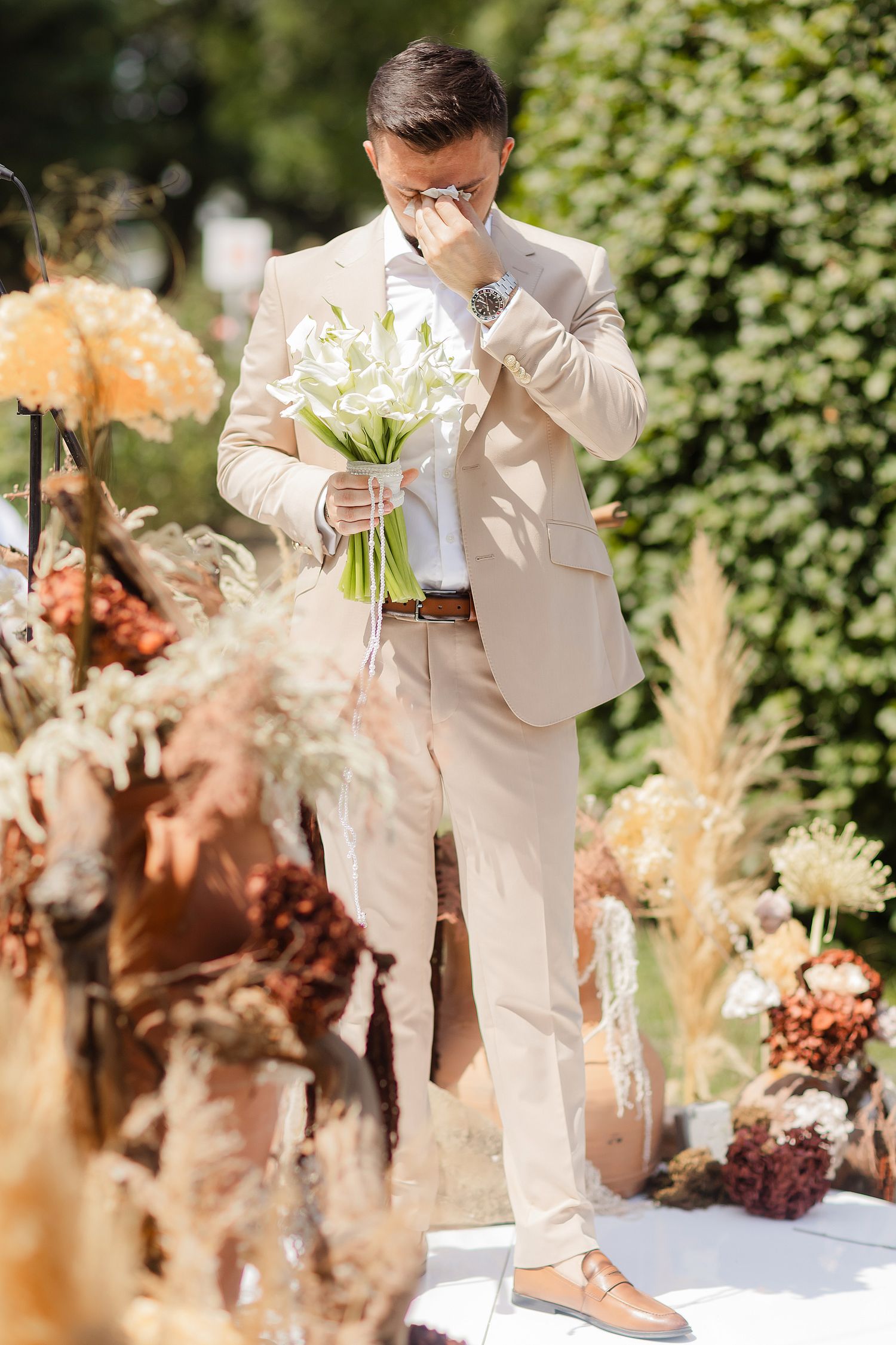 Emotional groom in beige suit holding white flower bouquet at outdoor wedding ceremony surrounded by dried floral arrangements.