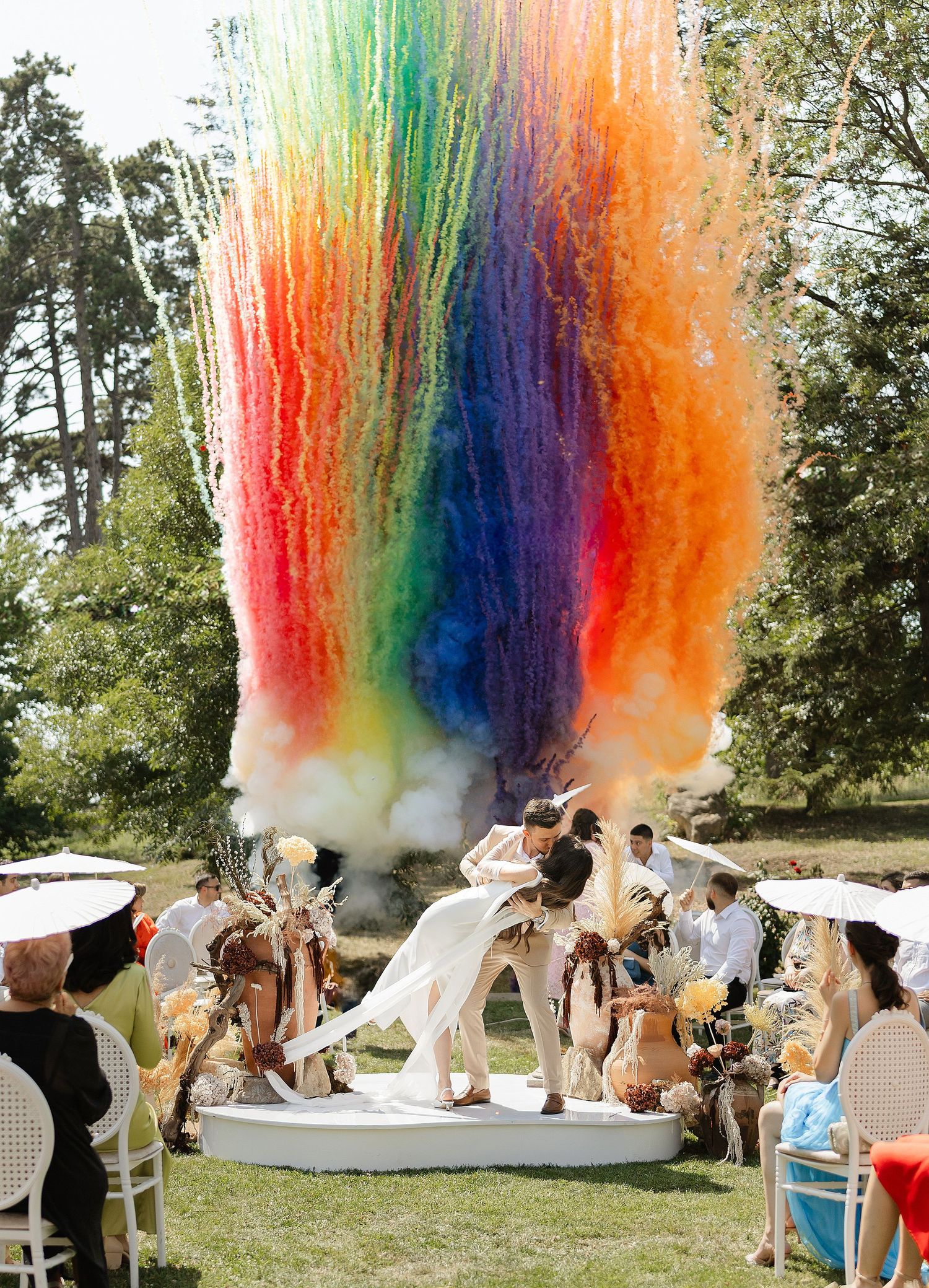 Rainbow smoke cannon explodes over couple kissing at outdoor wedding ceremony on white circular platform.