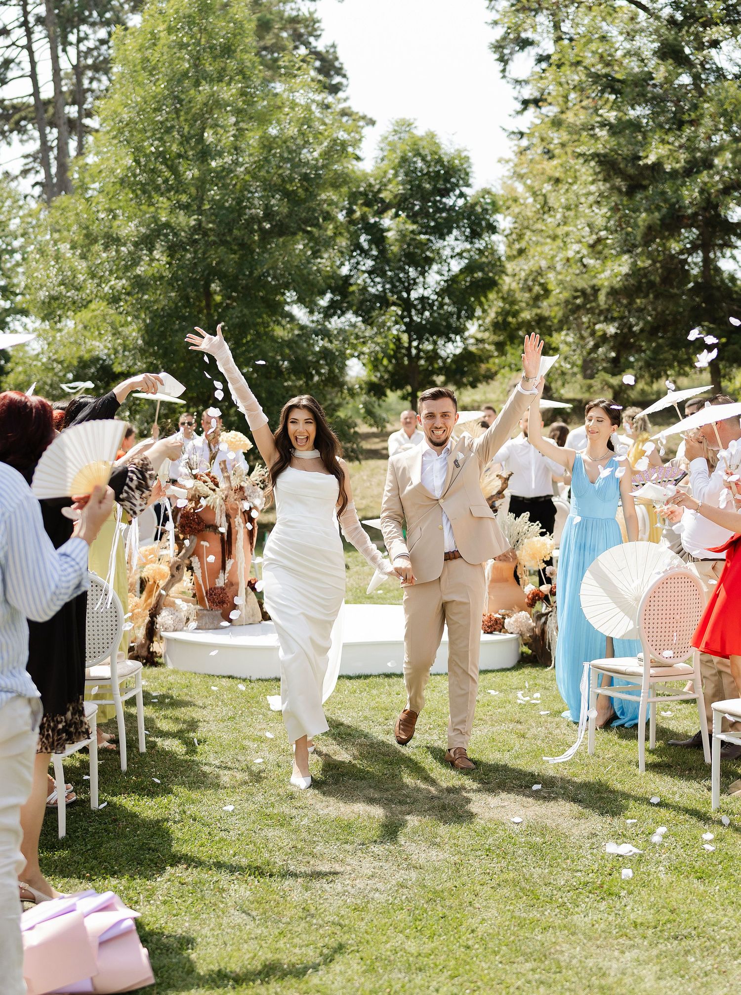 Joyful newlyweds walk down outdoor aisle as guests celebrate with confetti and fans on sunny wedding day.