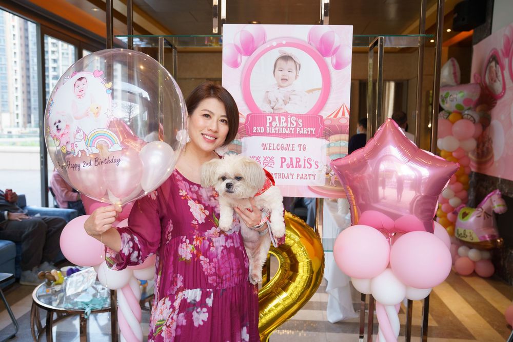 Woman in floral dress holding dog and balloons at Paris's birthday party with pink decorations.