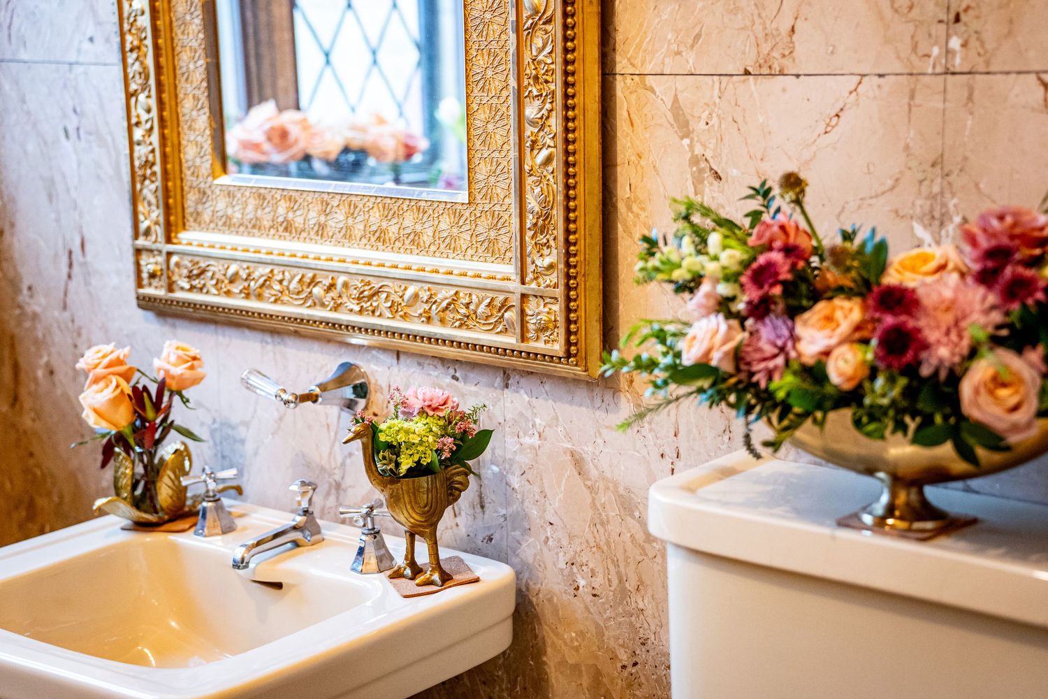 Elegant bathroom with gold-framed mirror, marble walls, white sink, and fresh floral arrangements in ornate vases.
