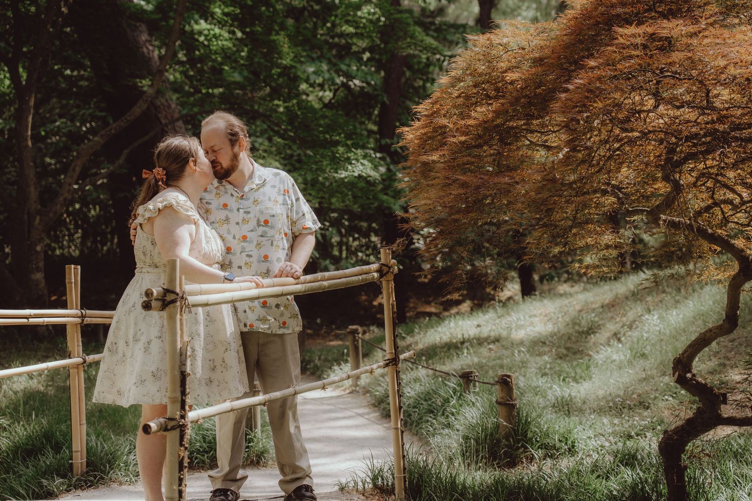 Couple in floral outfits walking on a garden path near a white bridge surrounded by lush green trees.