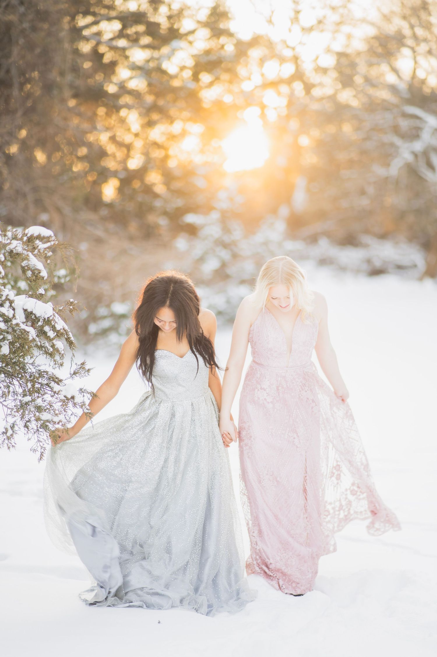 Two women in flowing gray and pink gowns walk hand-in-hand through a snowy forest at golden hour.