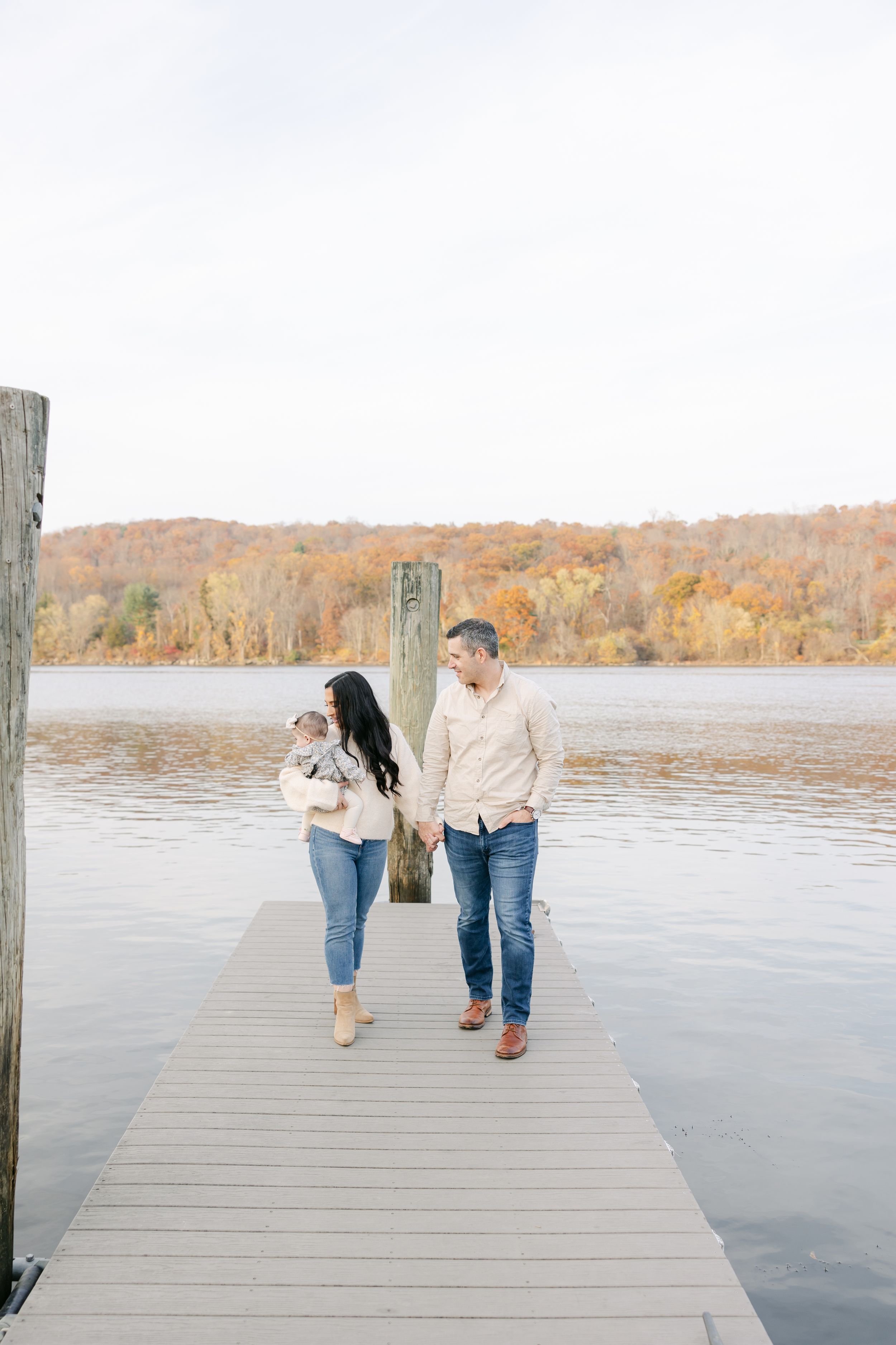 A couple holding hands on a dock, carrying a baby, with fall foliage and a calm lake in the background.