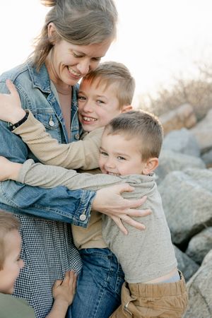 A smiling mother in a denim jacket hugs two happy young boys outdoors near rocky terrain.