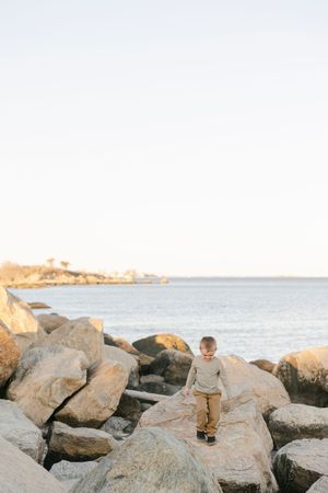 Young boy climbing large coastal rocks by the ocean at golden hour, wearing a beige sweater and tan pants.