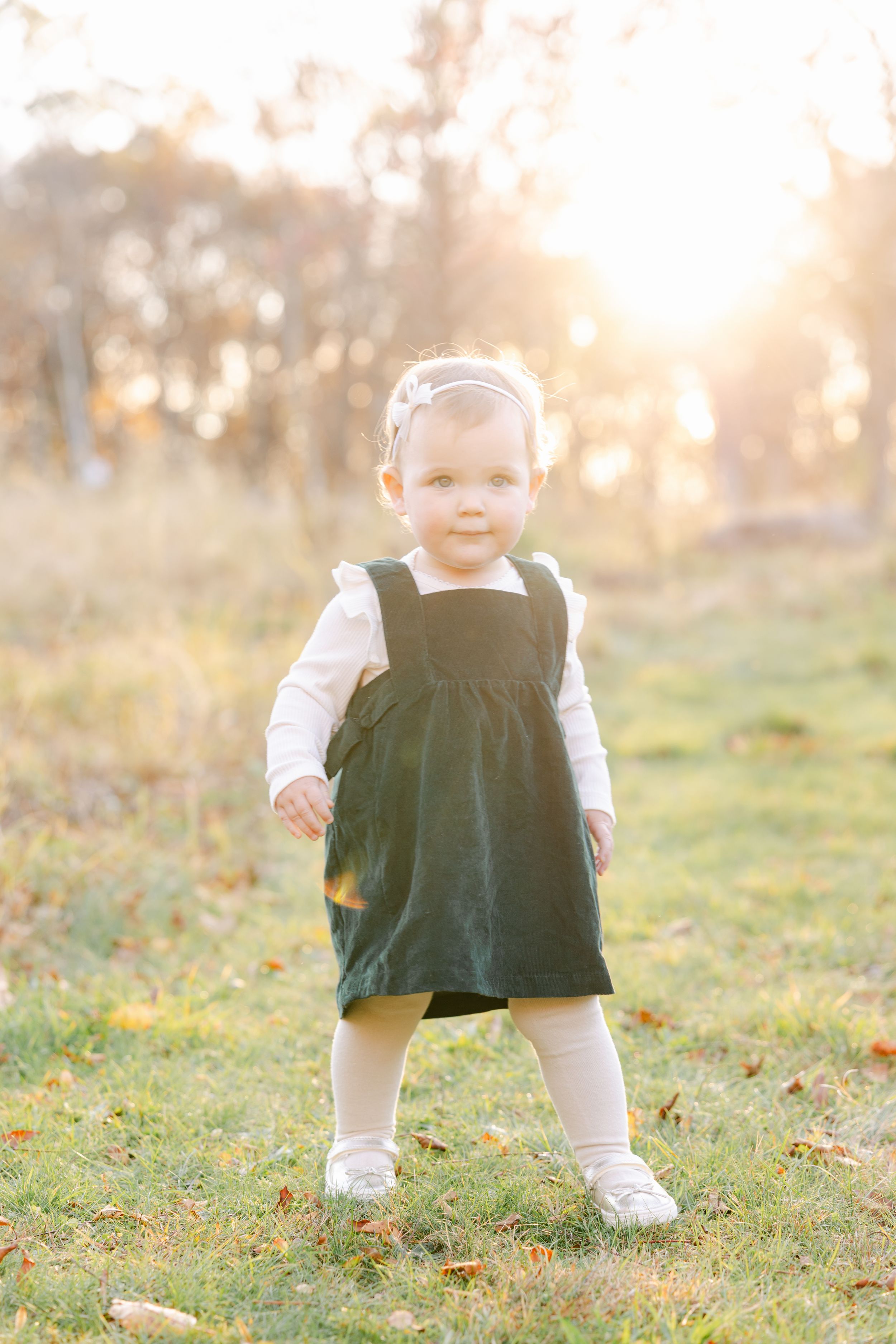 Toddler girl in dark green dress and white shirt standing on grass in golden sunset light outdoors.
