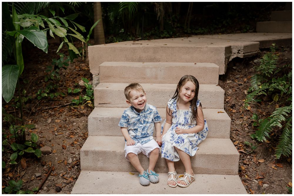 Two young siblings sitting together on stone steps smiling in a tropical garden setting.