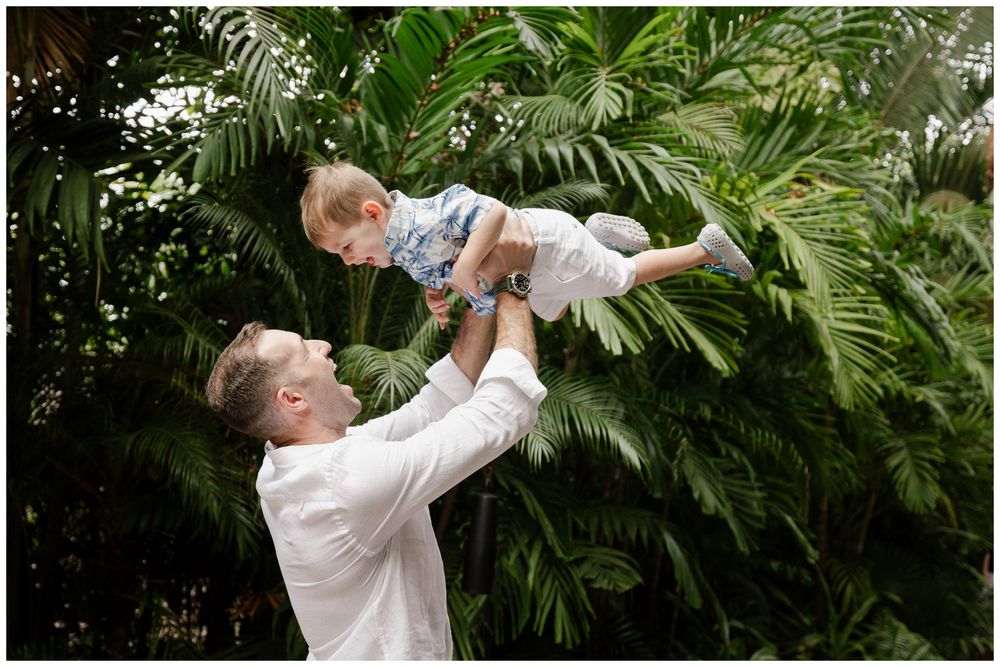 Father joyfully lifting smiling toddler high in the air surrounded by lush tropical greenery.