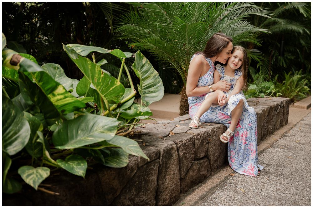 Mother kissing daughter while sitting on stone wall surrounded by tropical plants.