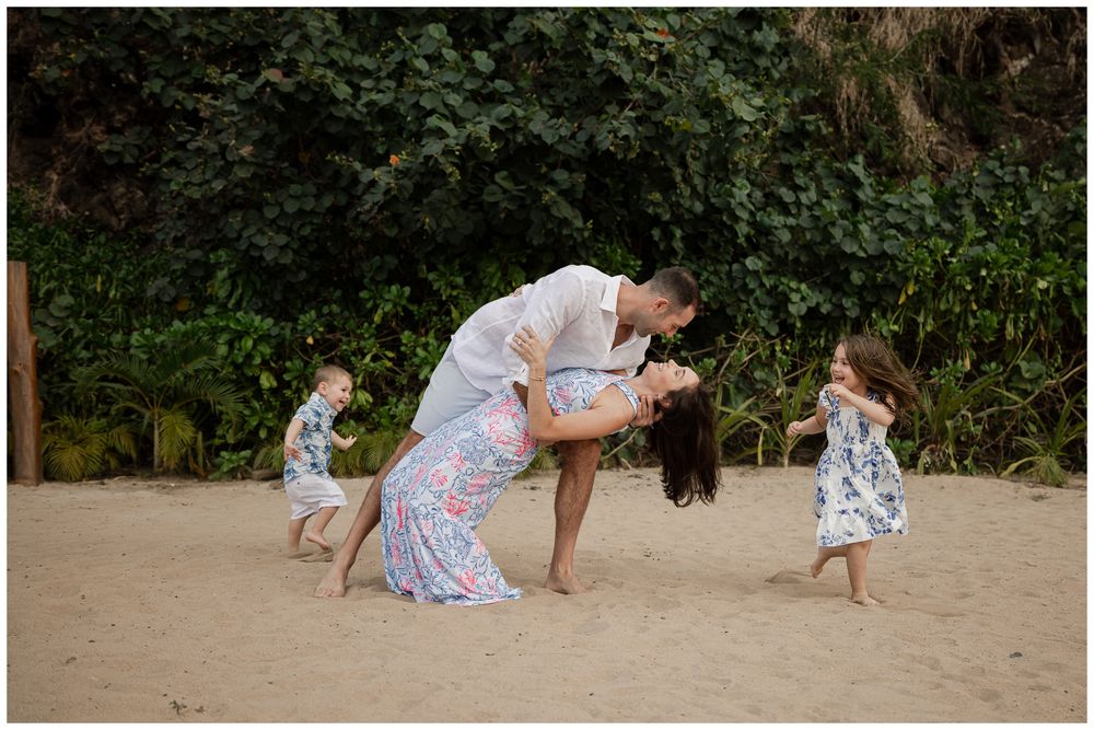 Couple sharing romantic dip kiss on beach while children play around them.