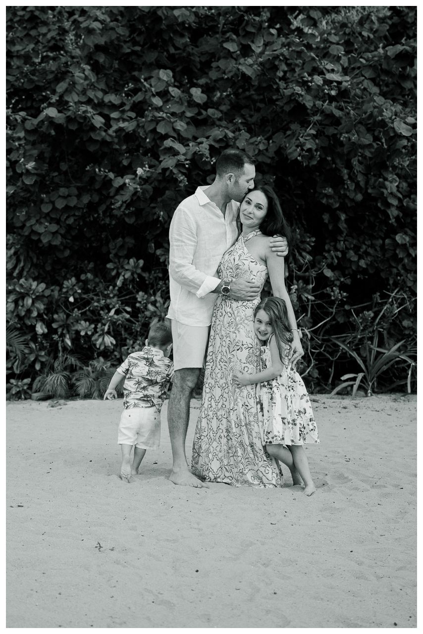 Black and white family portrait of parents with two young children embracing on a sandy beach.