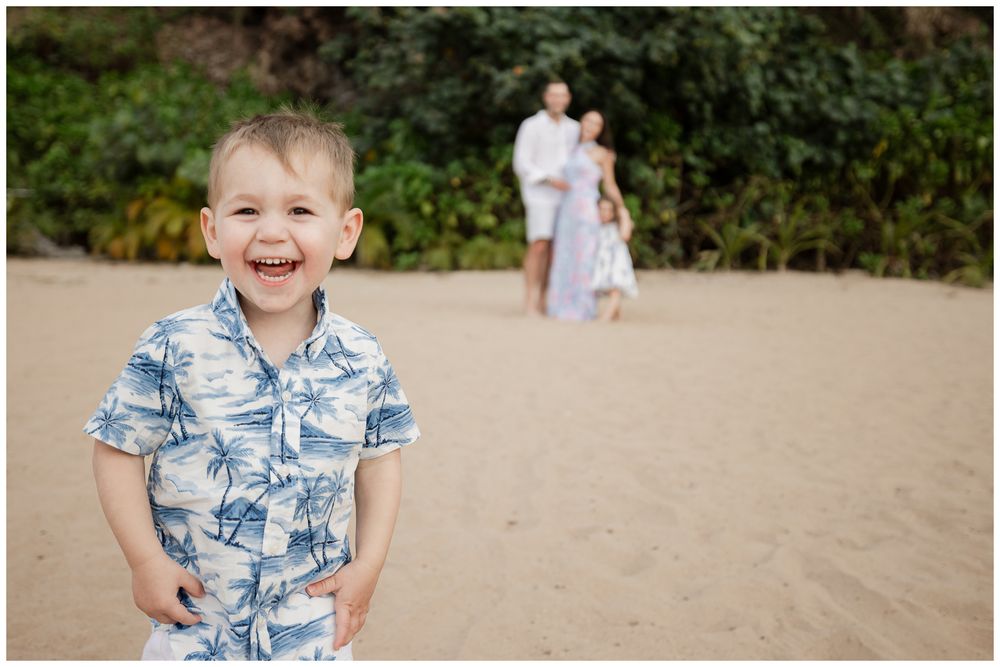A joyful toddler boy in a blue Hawaiian shirt laughs on a sandy beach with family in the background.