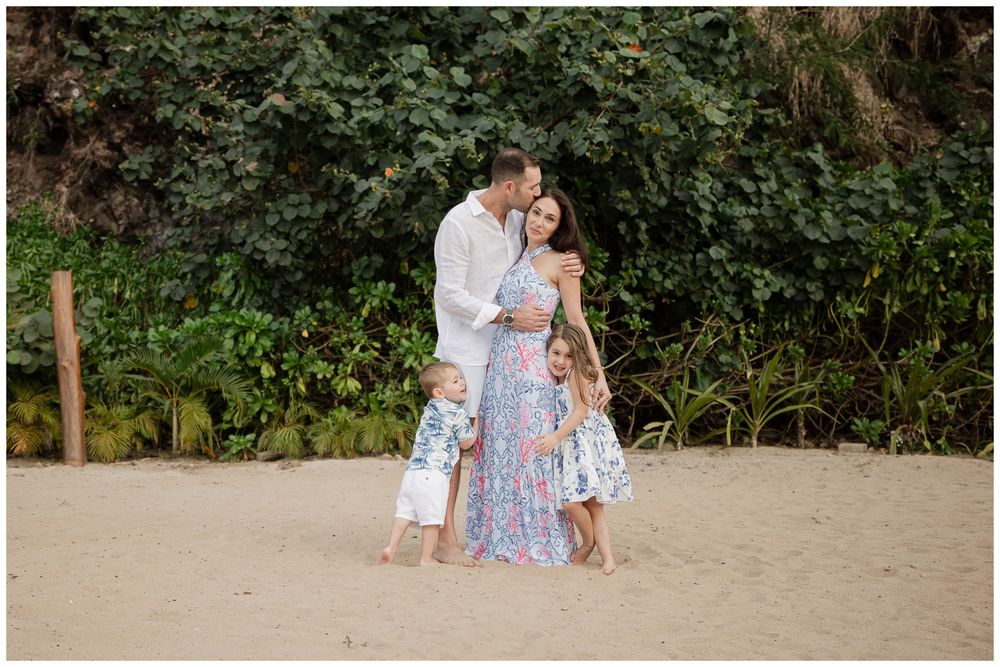 Family of four posing on sandy beach with lush green trees, parents embracing two young children in coordinating outfits.