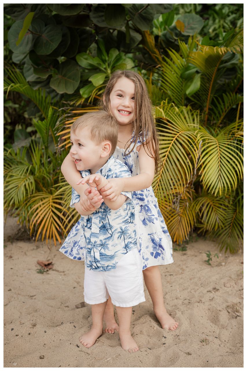 Two young siblings hug and smile on a sandy beach surrounded by lush tropical greenery in Hawaii.
