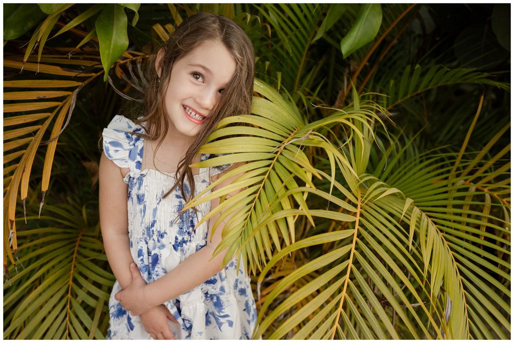 Young girl in floral dress smiling among lush tropical palm leaves in a vibrant garden setting.