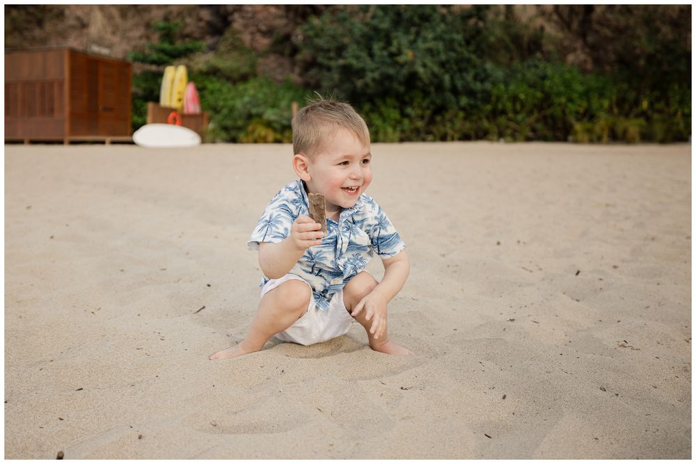Young boy in floral shirt laughing and playing in sand at beach.