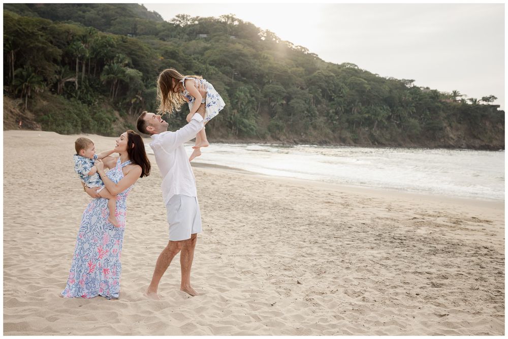 Family of four enjoying a golden hour beach photoshoot, parents lifting children on sandy shore with lush green cliffs behind.
