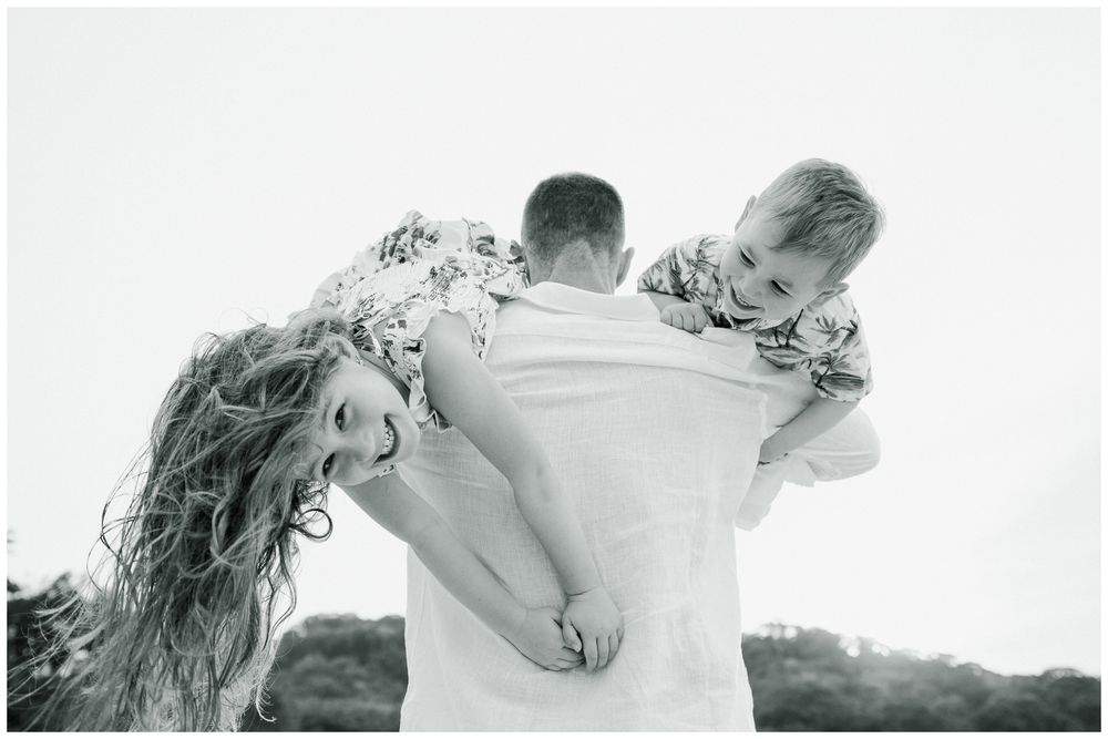 Black and white photo of father carrying two children on his shoulders outdoors.