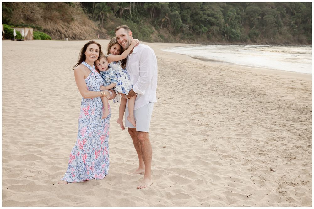 Family of four posing on sandy beach, parents holding young children, wearing casual summer outfits with ocean backdrop.