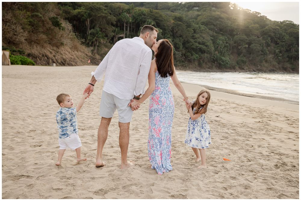 Family of four holding hands on a sandy beach at sunset, with lush green cliffs in the background.