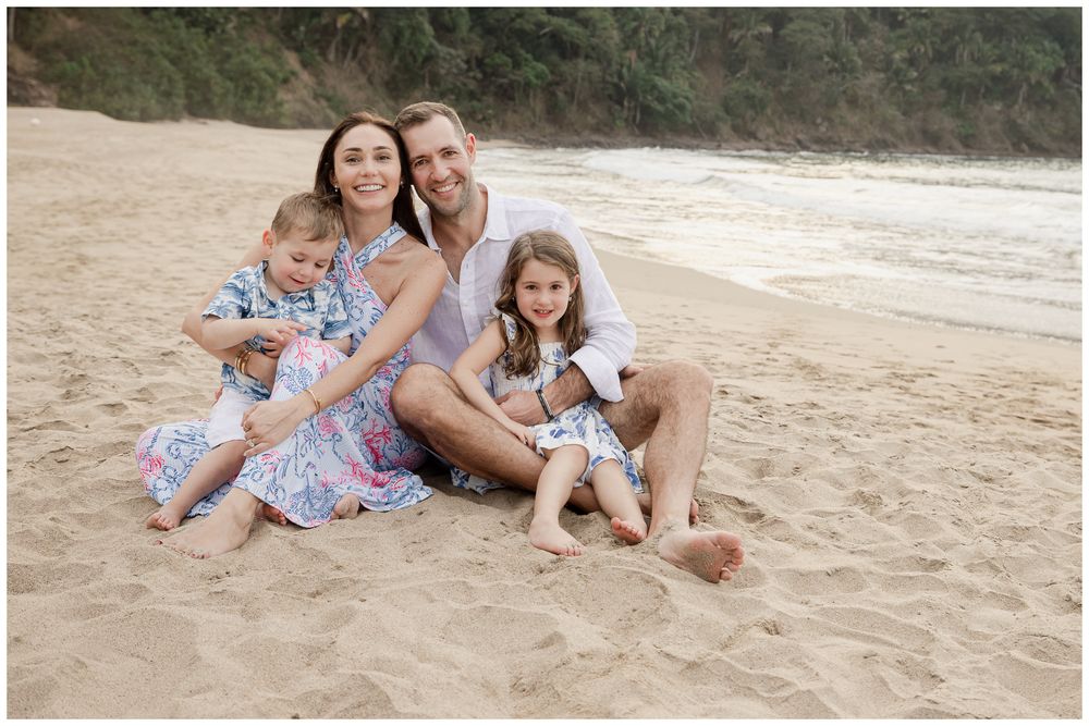 Family of four smiling together on sandy beach, parents and two young children in coordinating outfits.