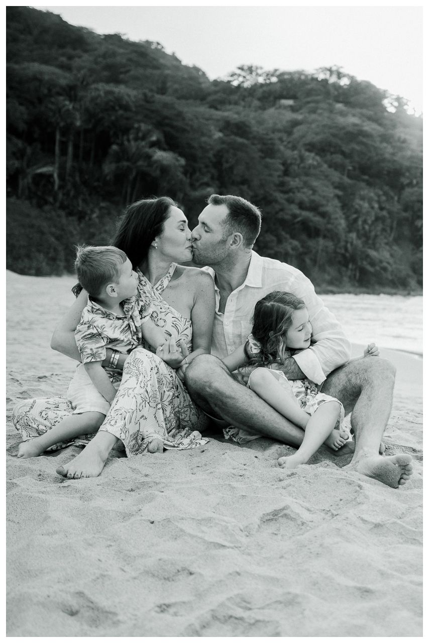 Black and white beach photo of a couple kissing while two young children cuddle beside them on sandy shore.