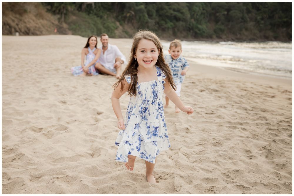 Young girl in blue floral dress running on sandy beach with family sitting behind her.