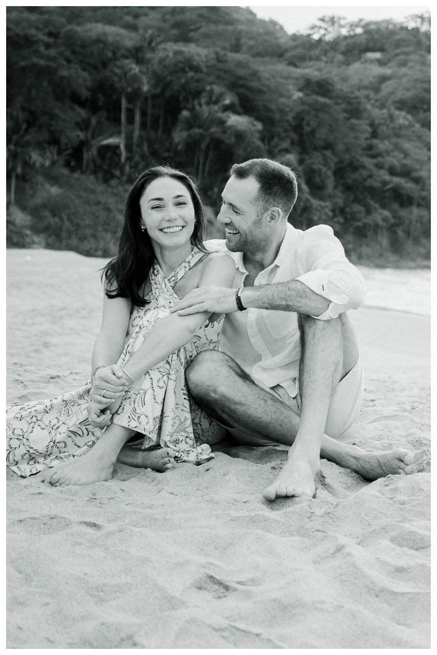 Black and white photo of a smiling couple sitting together on a sandy beach with lush trees in the background.