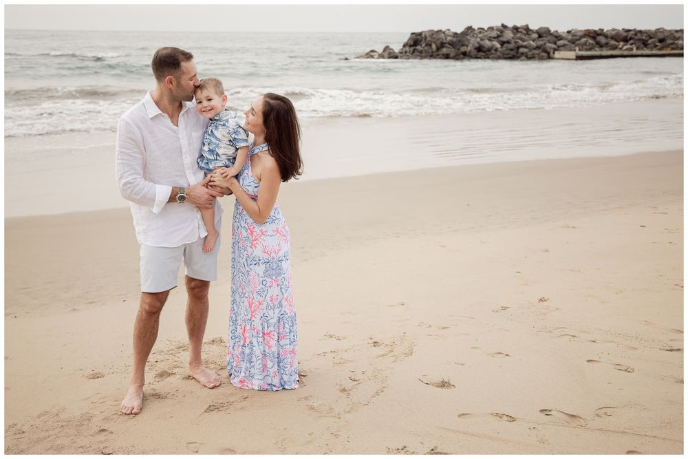 A young family with a baby poses on a sandy beach, with ocean waves and rocky jetty in the background.