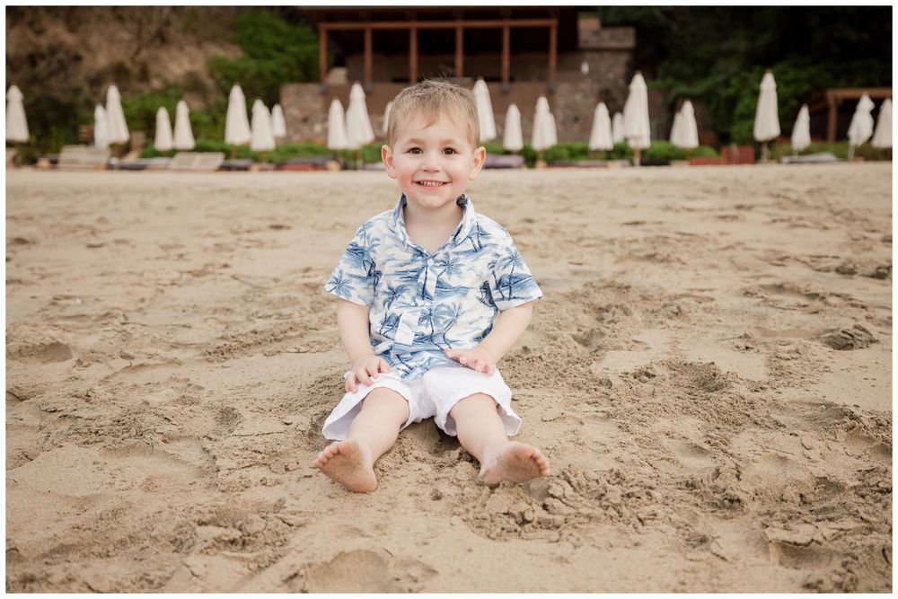 Adorable toddler boy in Hawaiian shirt sitting and smiling on sandy beach.